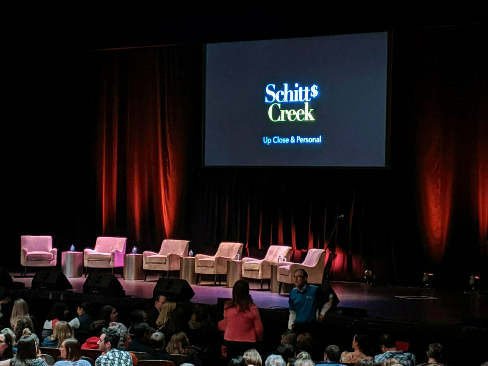 A stage is set with chairs and a large screen displaying Schitt's Creek: Up Close & Personal in front of an audience.