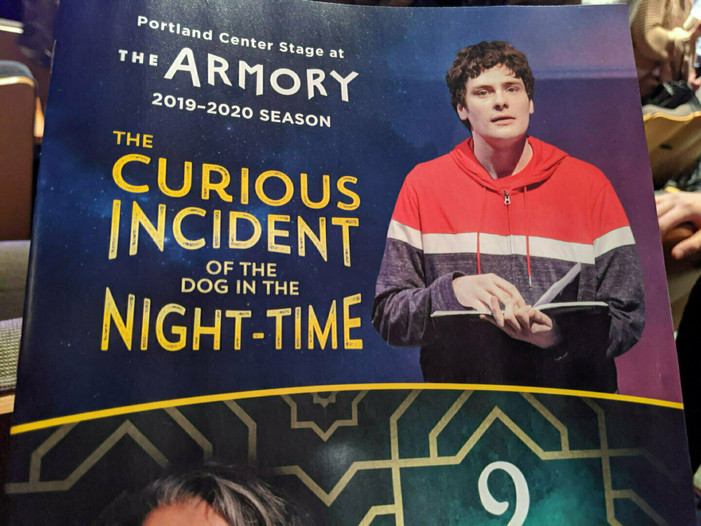 A theater program cover for The Curious Incident of the Dog in the Night-Time at Portland Center Stage at The Armory features a person in a red and white jacket.