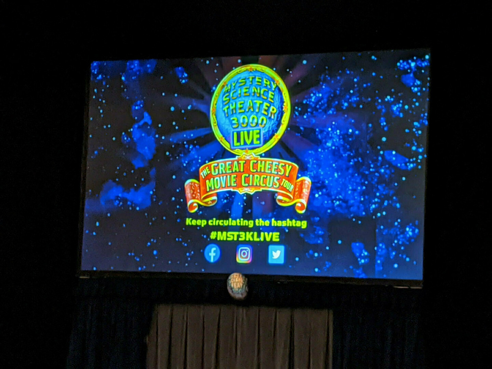 A promotional screen for Mystery Science Theater 3000 Live: The Great Cheesy Movie Circus Tour features social media handles and the hashtag #MST3KLIVE.