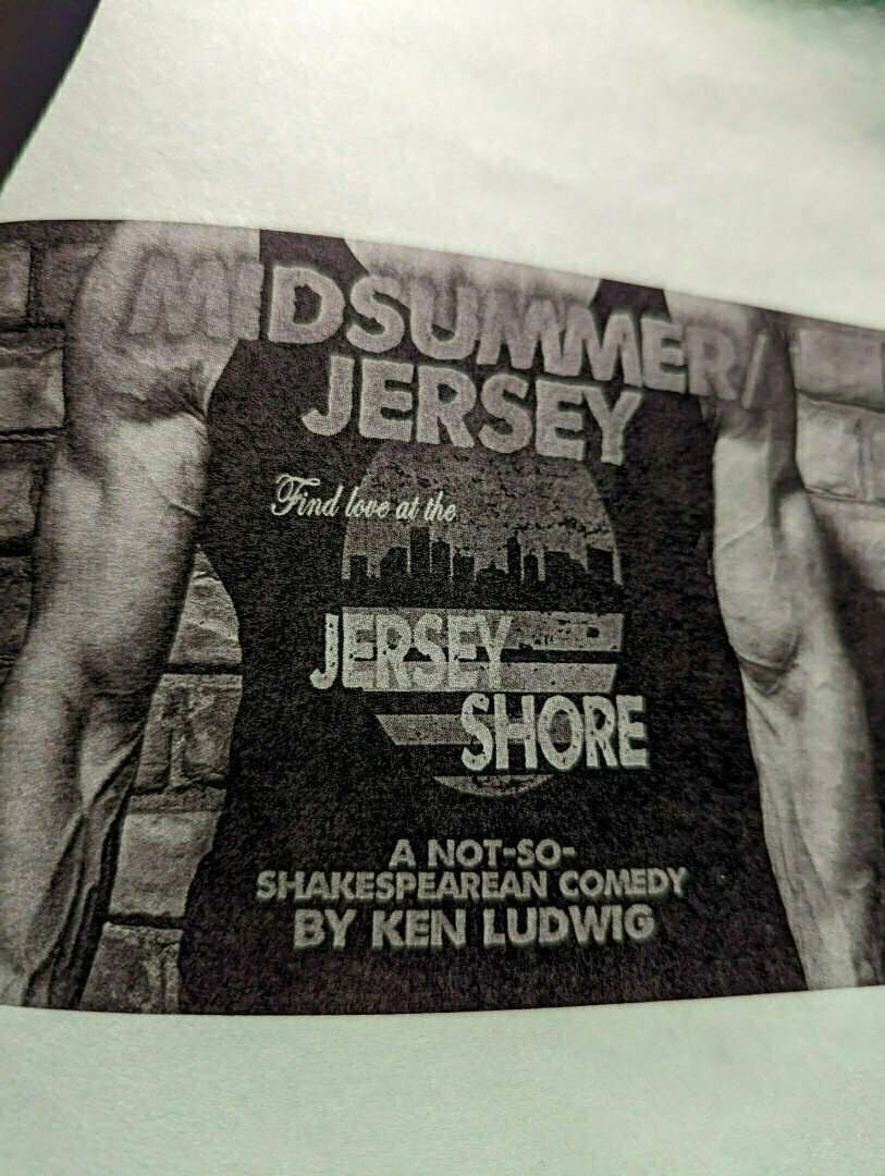 A muscular person in a sleeveless top is standing in front of a brick wall, and the shirt has text promoting a comedic play titled Midsummer/Jersey by Ken Ludwig.