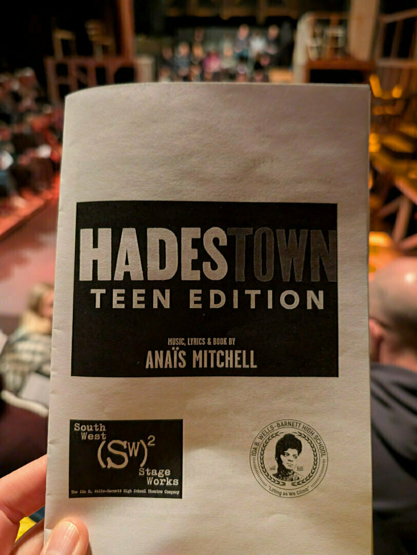 A person is holding a program for Hadestown Teen Edition with a theater audience and stage in the background.