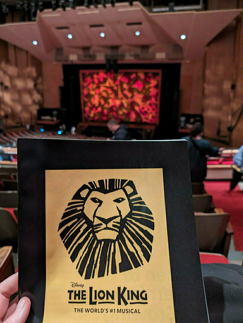 A theatergoer holds a program for The Lion King musical, with the stage in the background.