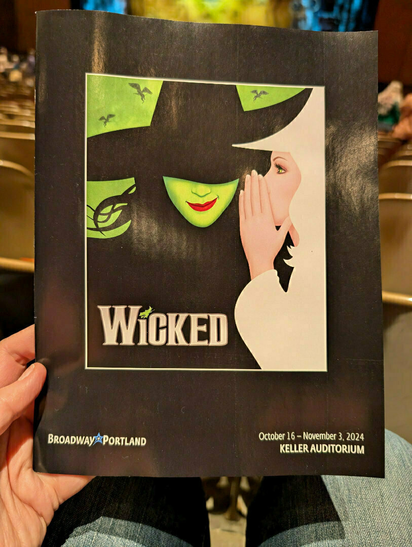 A playbill for the musical Wicked is being held, showing a stylized illustration of a green-skinned witch and another figure whispering.