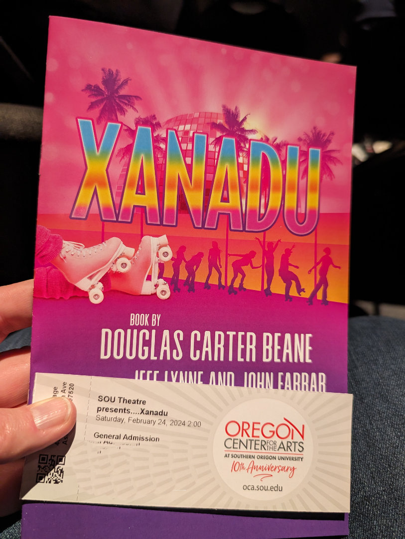 A person is holding a colorful theater program for Xanadu along with an admission ticket for a performance at the Oregon Center for the Arts.