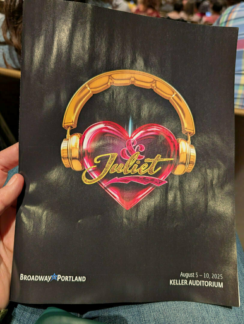 A person is holding a Broadway in Portland playbill for & Juliet featuring a heart with headphones, set to run at Keller Auditorium from August 5-10, 2025.