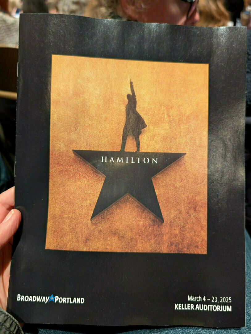 A hand holds a playbill for the musical Hamilton, scheduled to be performed at the Keller Auditorium in Portland from March 4-23, 2025.