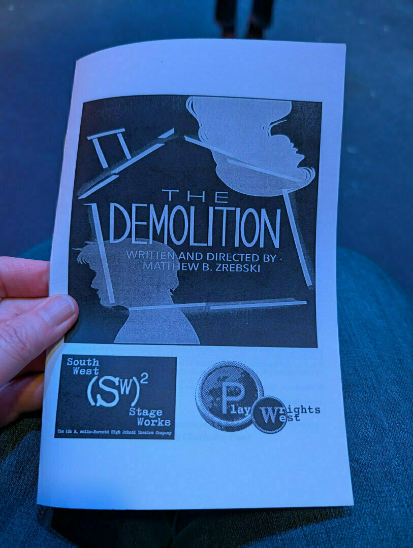 A person is holding a theater program titled The Demolition, written and directed by Matthew B. Zrebski, featuring silhouettes and logos for South West Stage Works and Playwrights West.