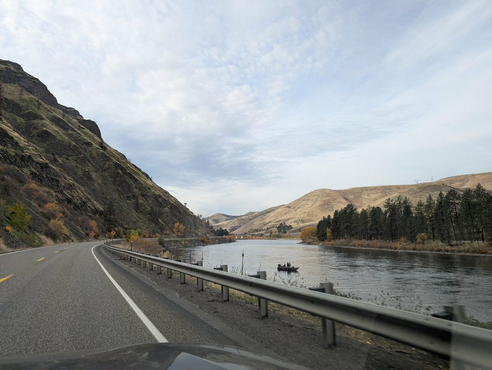 A scenic road winds alongside a river with hills and trees under a cloudy sky.