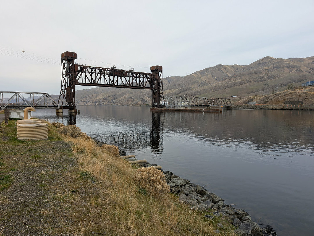 A railroad lift bridge spans across a calm river surrounded by hills and grassy terrain.