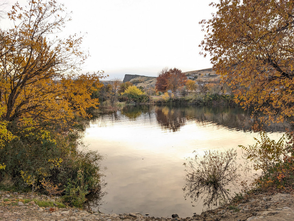A serene lake is surrounded by trees with autumn foliage and rocky terrain, reflecting a tranquil landscape.