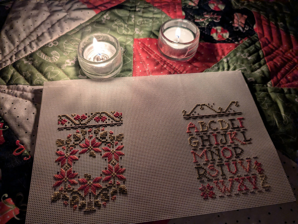 A piece of cross-stitch embroidery featuring decorative patterns and the alphabet is set on a quilted surface with two lit candles nearby.