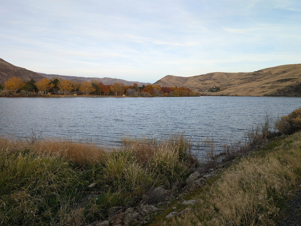 A serene lake is surrounded by grassy banks and distant hills under a partly cloudy sky.