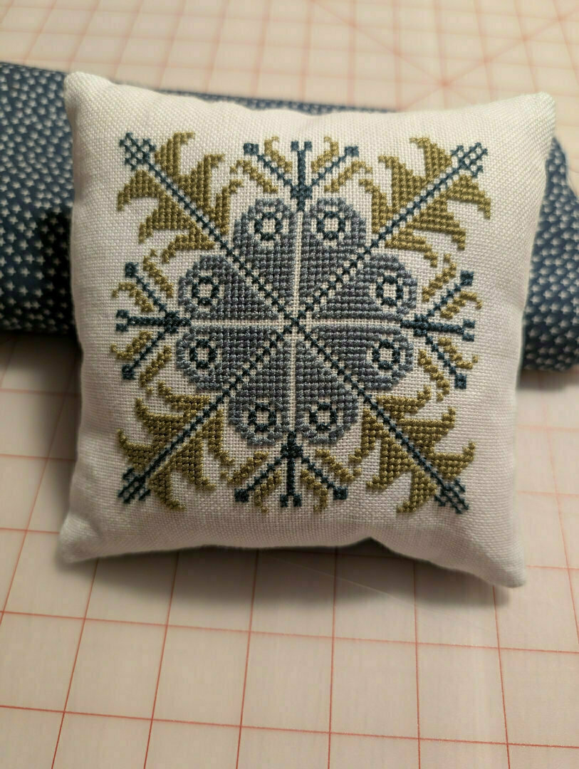 A small pillow features an intricate cross-stitched pattern in blue and green, resting on a patterned fabric.