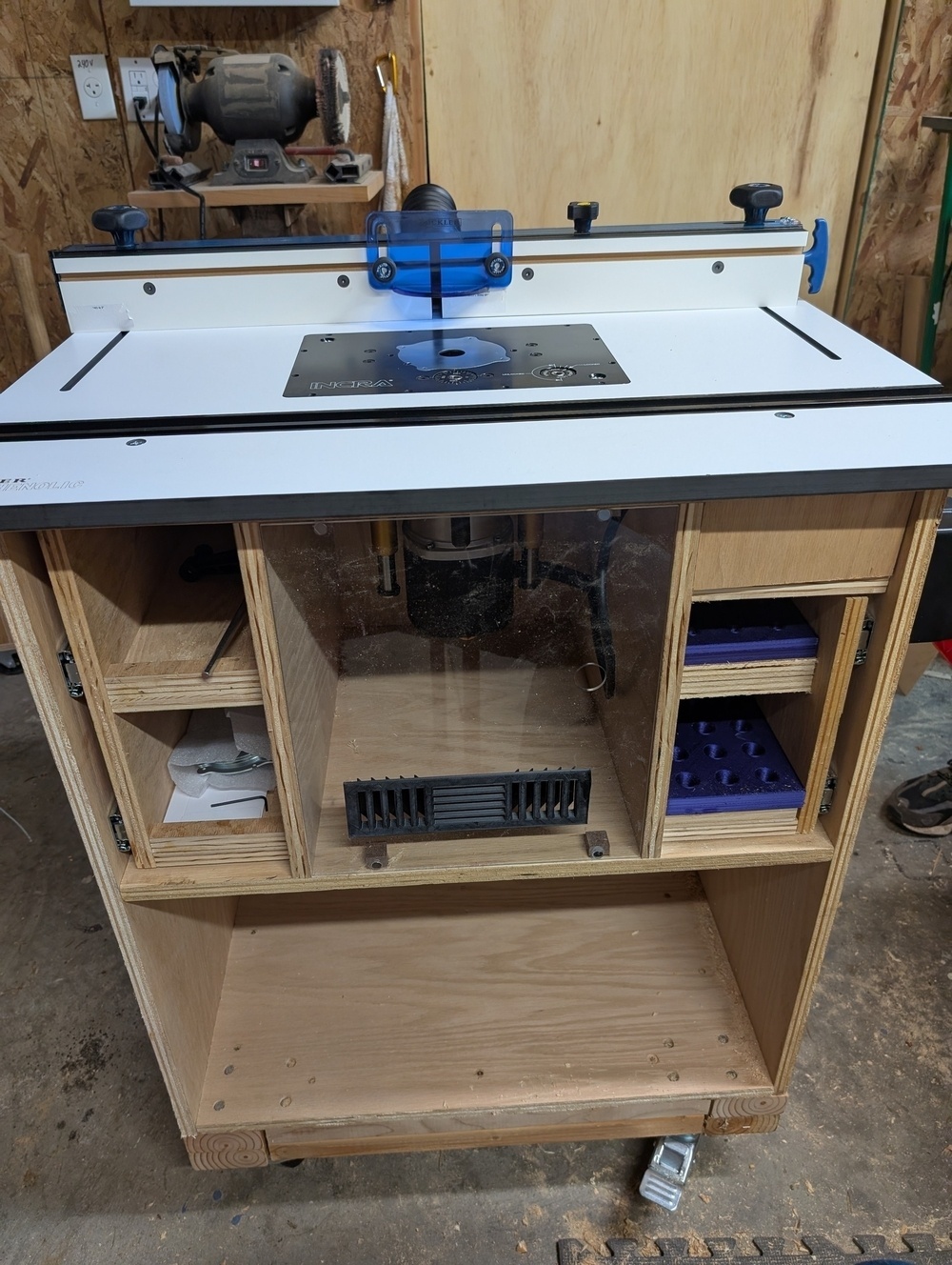 A woodworking router table is equipped with a blue router and storage shelves, located in a workshop.