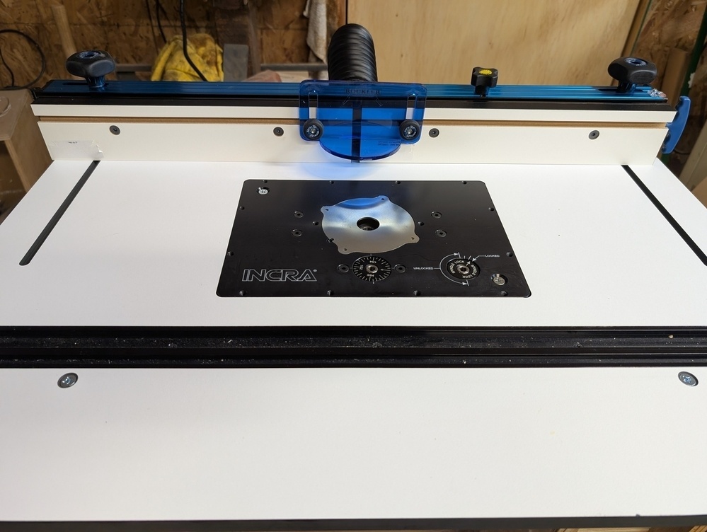 A woodworking router table setup with a white surface, an Incra router plate, and a blue fence on a workbench.