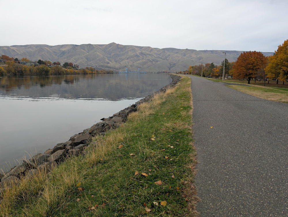 A paved pathway runs alongside a calm river with autumn trees and distant hills in the background.