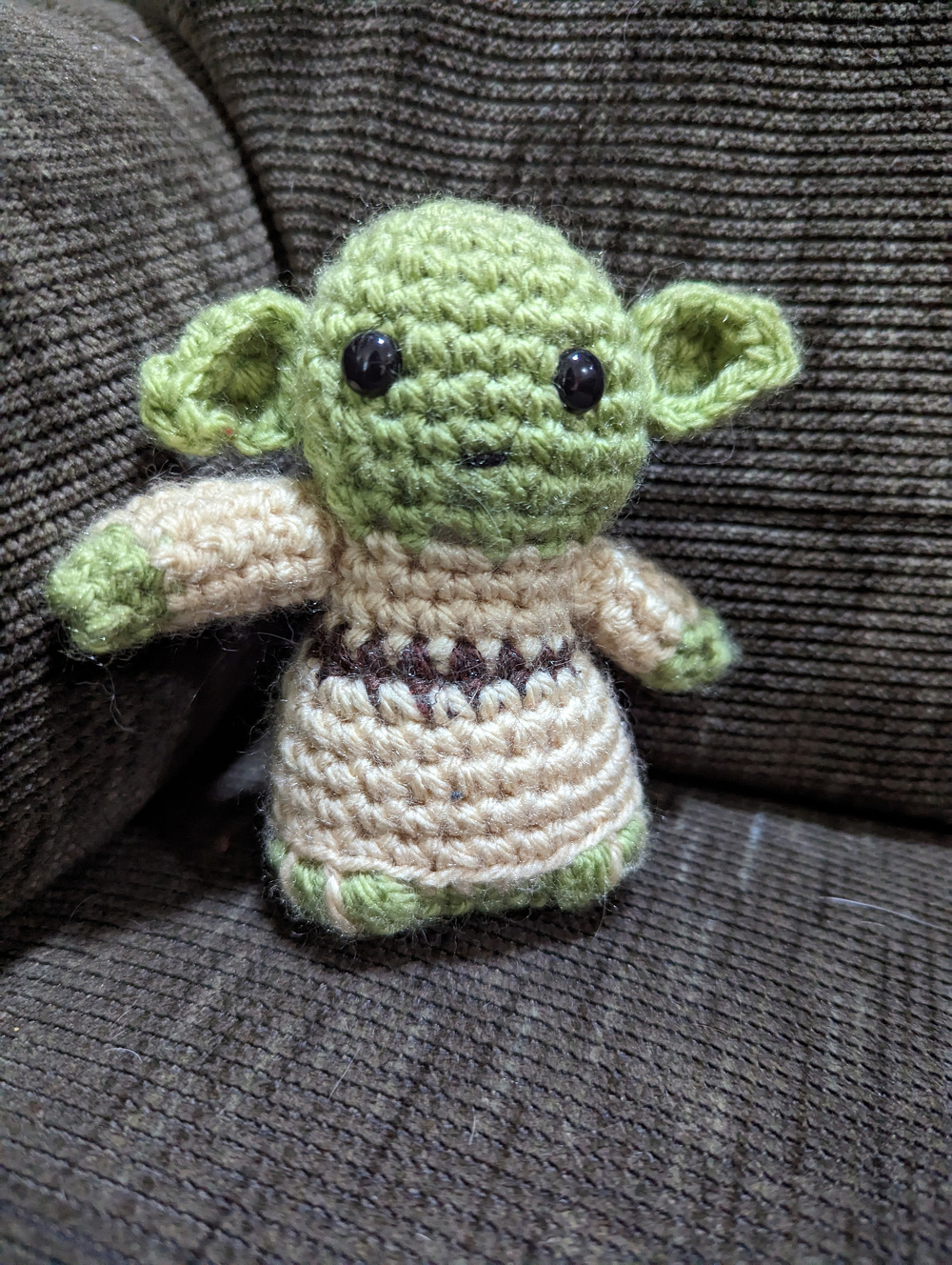 A crocheted doll resembling a popular fictional character with green skin and large ears is seated on a brown fabric surface.