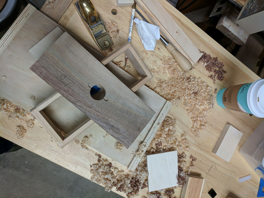A woodworking project is underway on a cluttered workbench with various tools, wood shavings, and a cup of coffee.