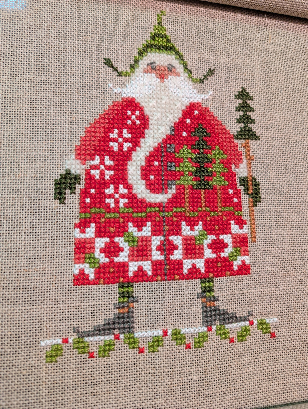 A cross-stitched design depicts a festive figure in a red coat adorned with Christmas trees, holding a small tree, and wearing a green hat.
