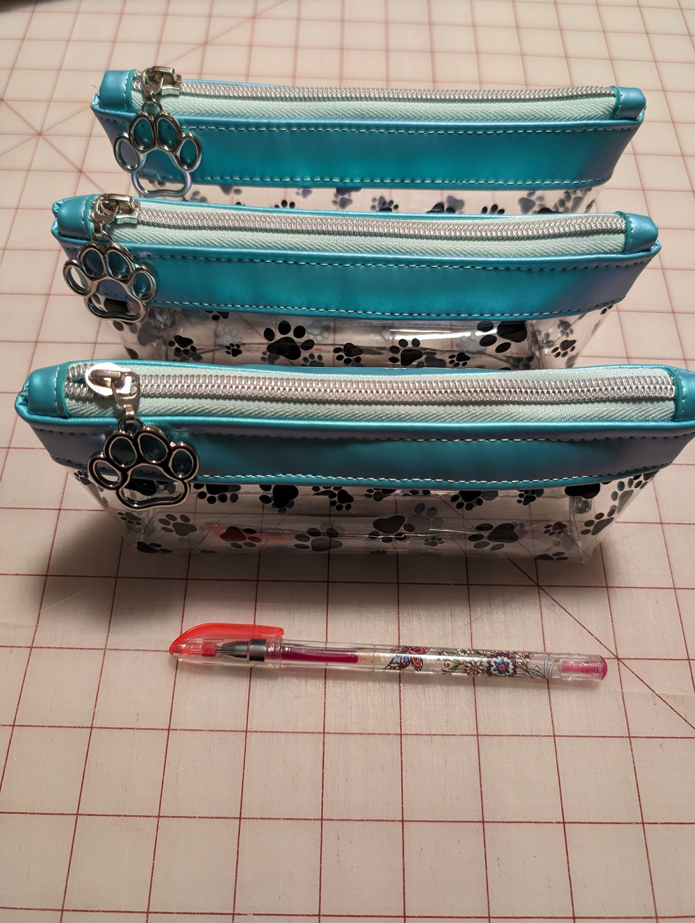 Three zipper pouches with paw print designs are lined up alongside a pen on a checkered surface.