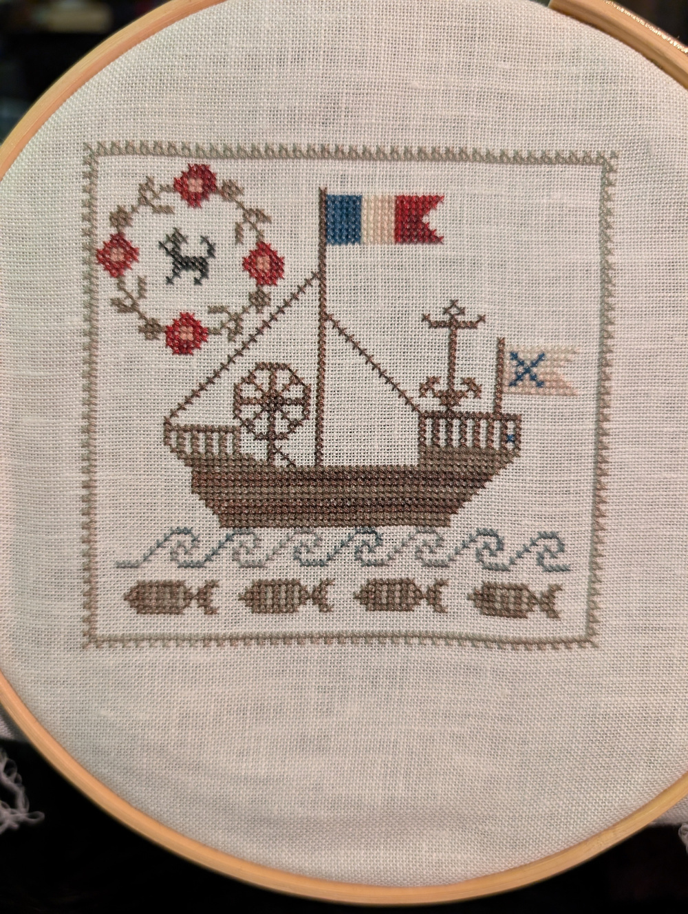 A cross-stitched tapestry shows a ship with a flag, fish below, and a decorative border including a floral pattern and an animal motif.