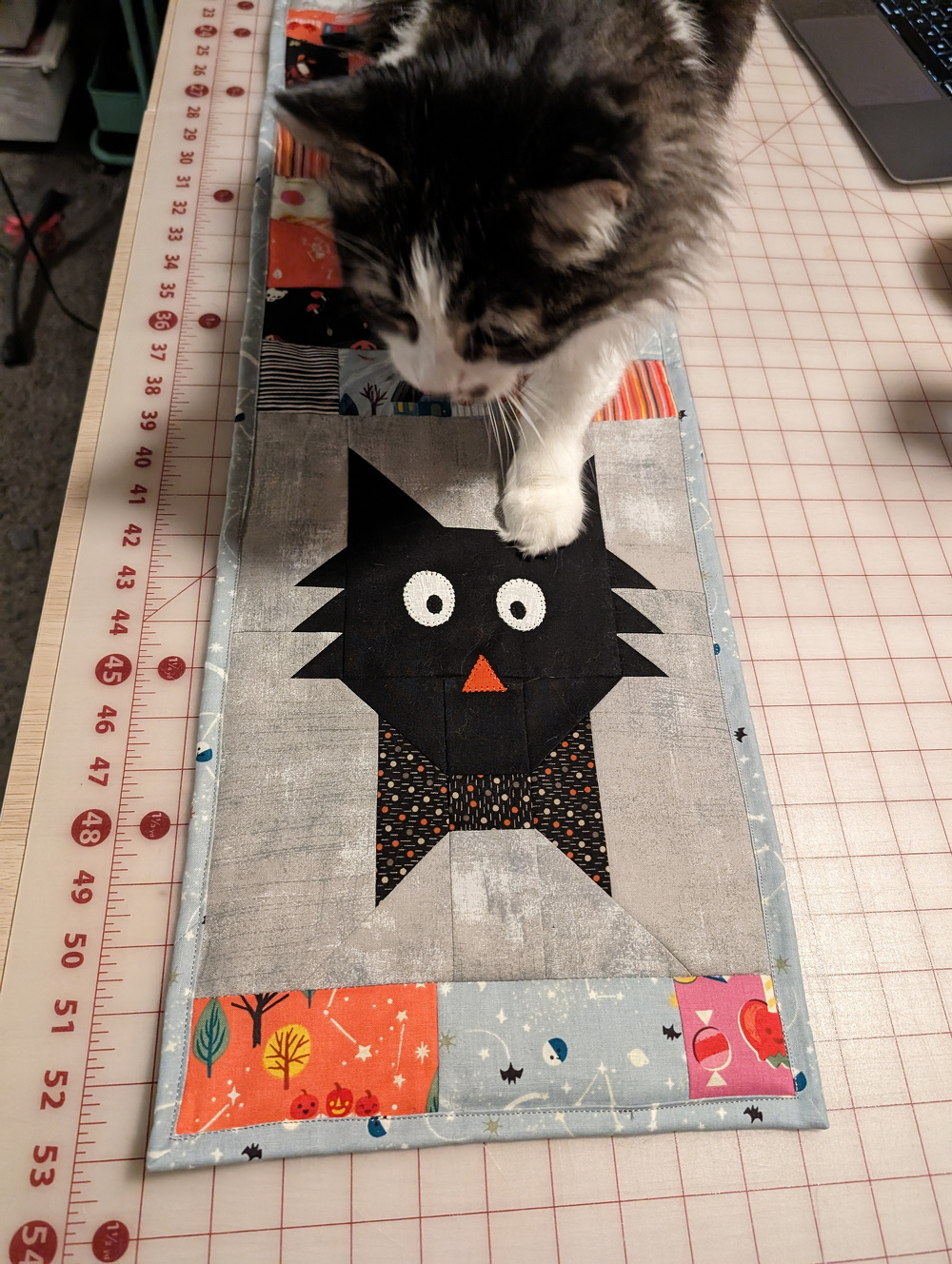A cat steps on a quilt featuring a patchwork design of a black cat with wide eyes and an orange nose.