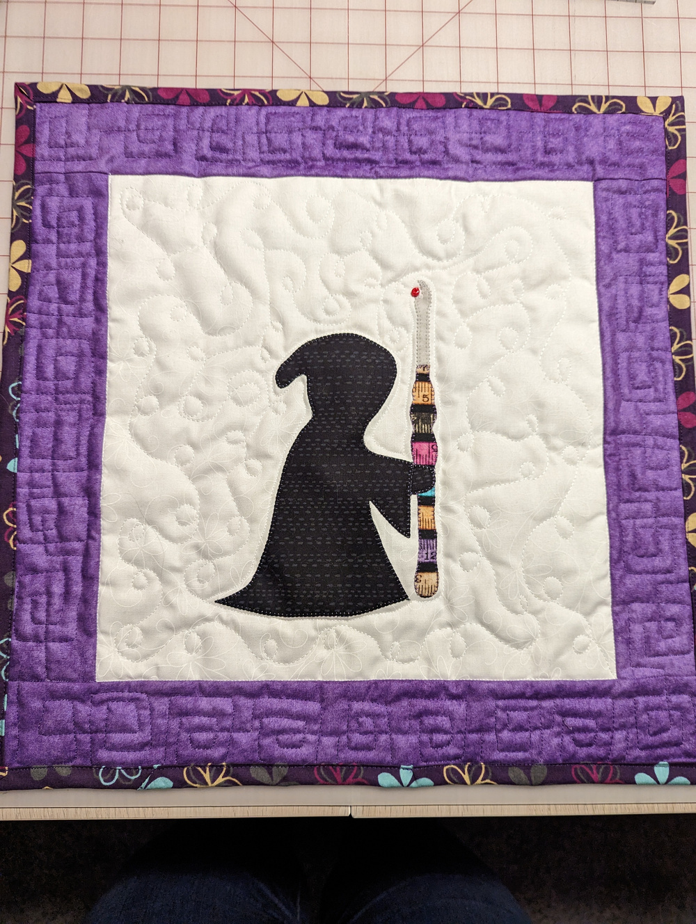 A quilted panel features a silhouette of a hooded figure holding a staff, surrounded by a purple geometric border with floral accents.