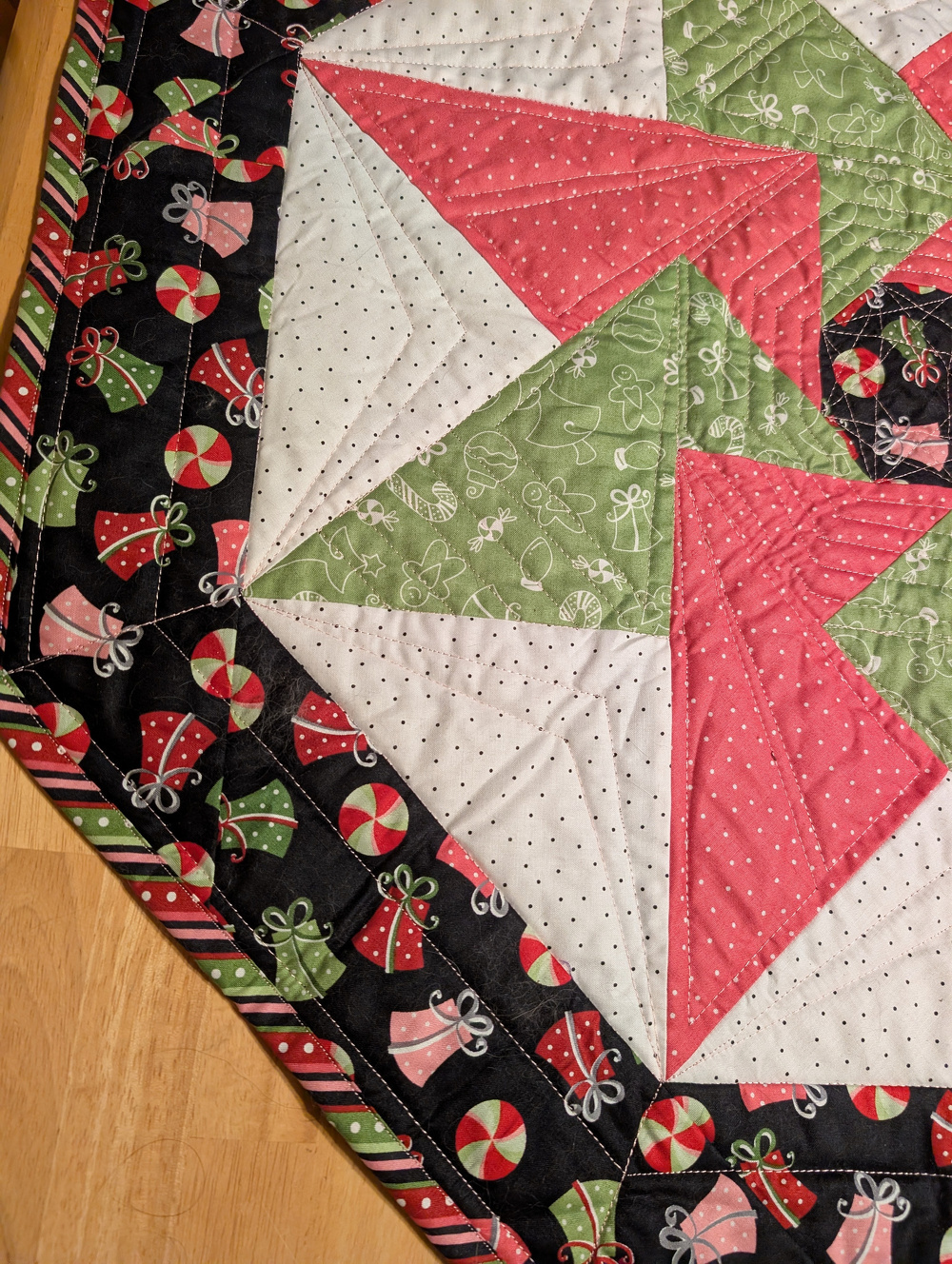 A quilt with a geometric pattern in red, green, and white features holiday-themed prints with bells and gifts on the border.