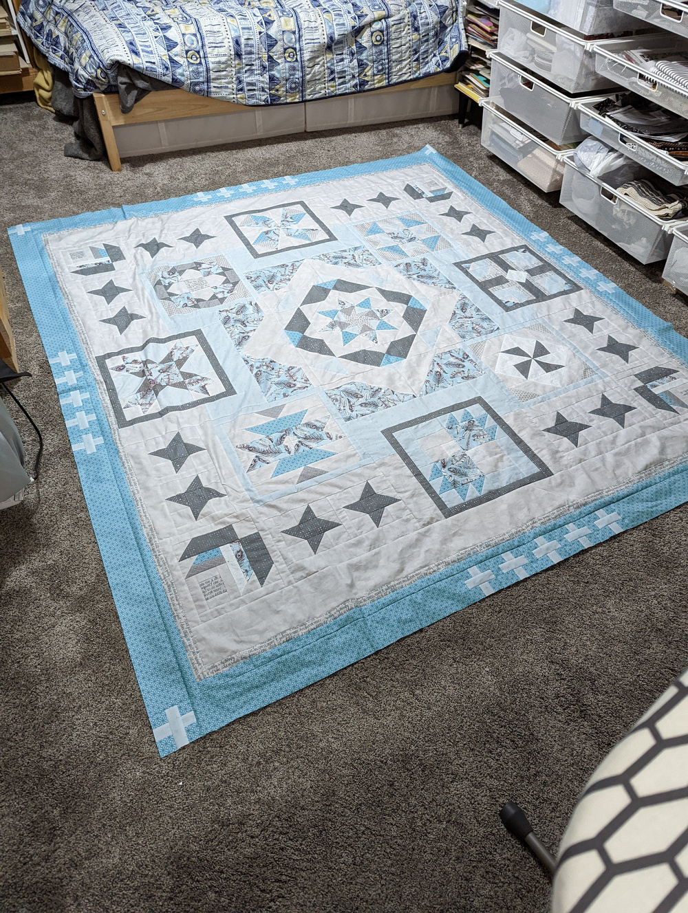 A quilt featuring a geometric pattern with shades of blue, gray, and white is laid out on the floor in a room with a carpeted surface and storage bins.