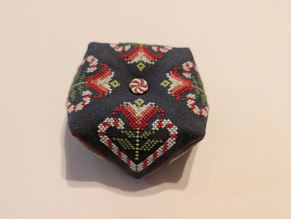 A decorative, hexagonal, fabric pincushion features intricate cross-stitched heart designs and a red and white button on top.