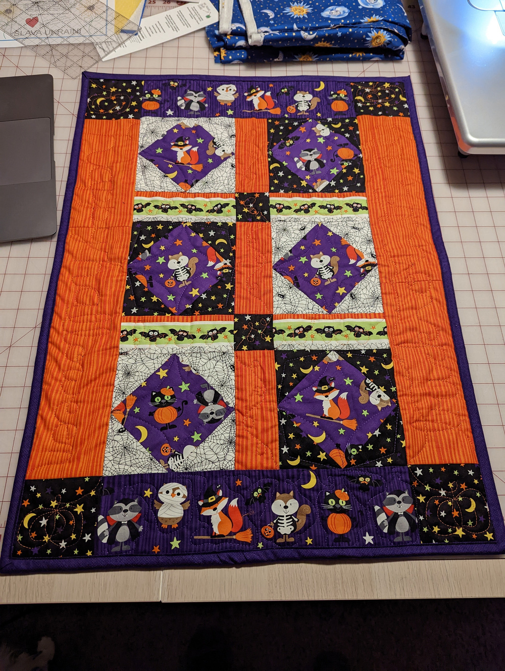 A colorful Halloween-themed quilt features cute animal characters in costumes, surrounded by patterns of stars, pumpkins, and bats.