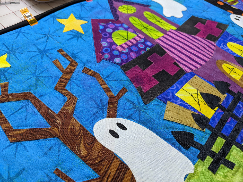 A colorful quilt features a whimsical haunted house scene with ghosts, stars, and a tree.