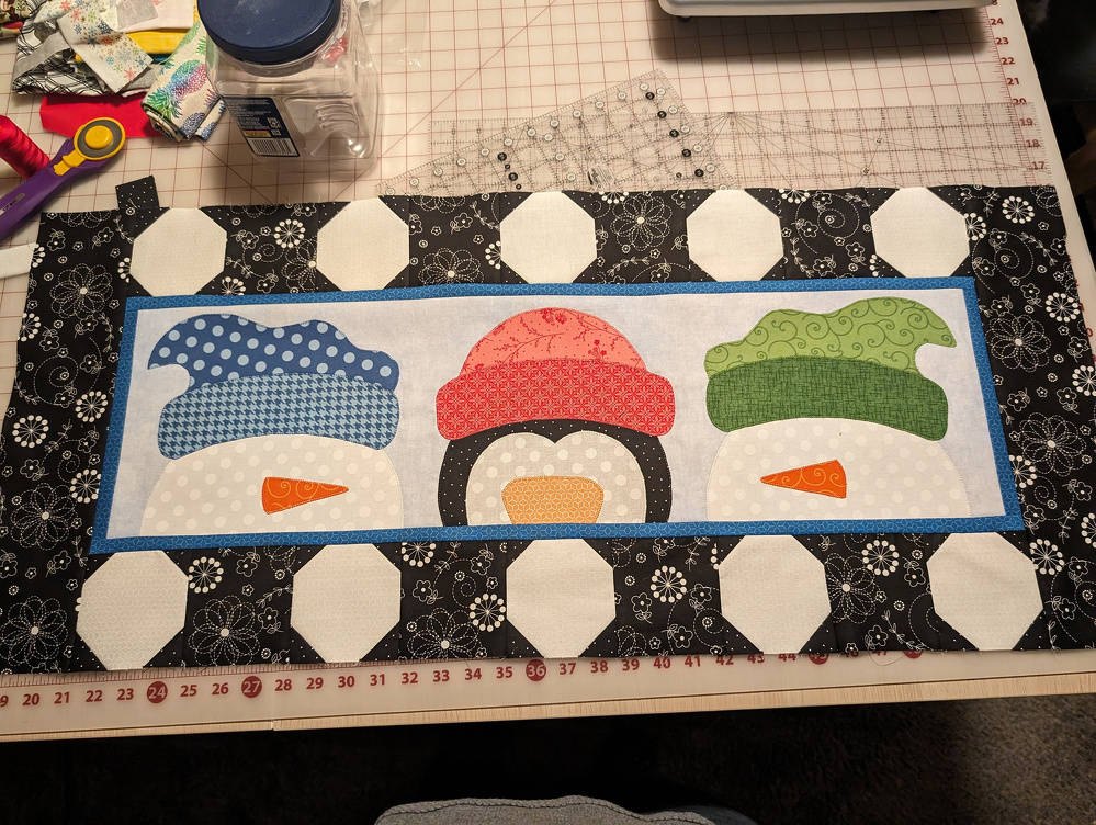 A quilted table runner features three stylized snowmen with colorful hats and orange noses against a patterned black and white background.