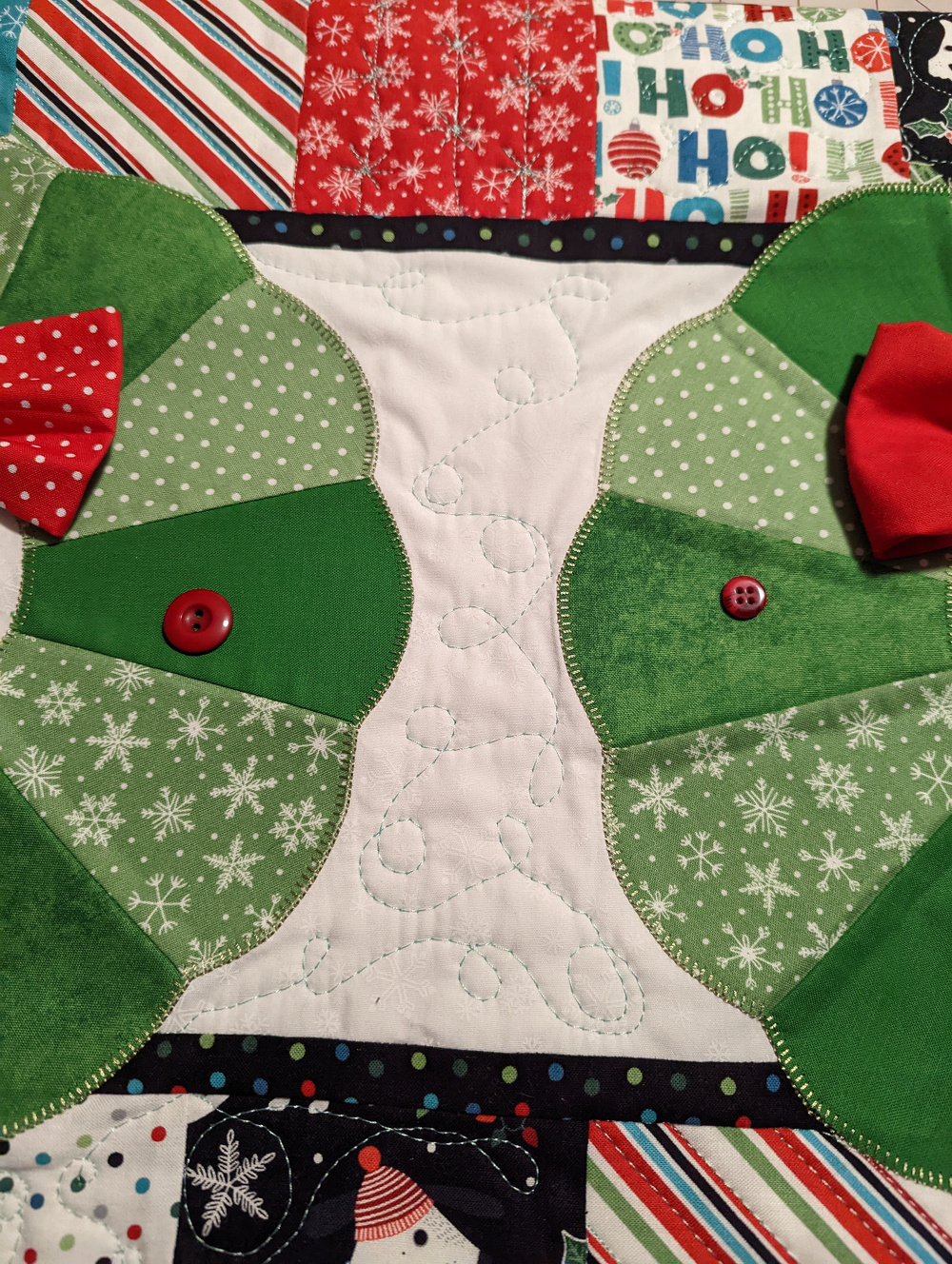 A colorful quilt features a festive holiday design with green and red patterned fabrics, red bow ties, and buttons.