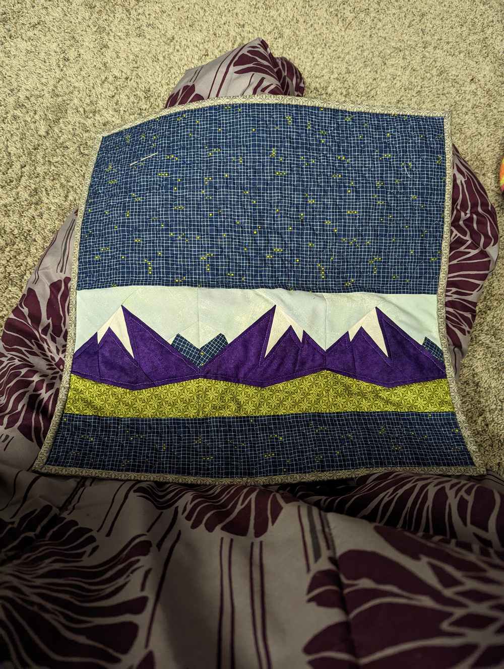 A small quilt square features a design of purple mountains, a green patterned strip, and a blue sky with star-like dots.
