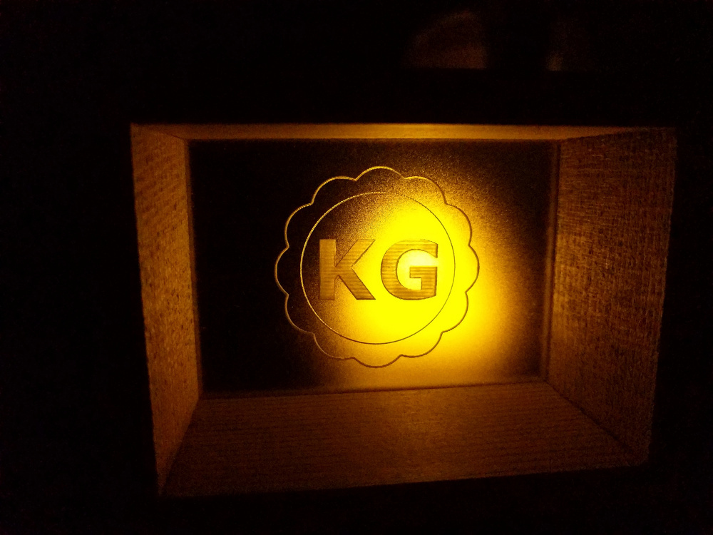 A warmly lit box displays the letters KG on a textured background with a decorative border.