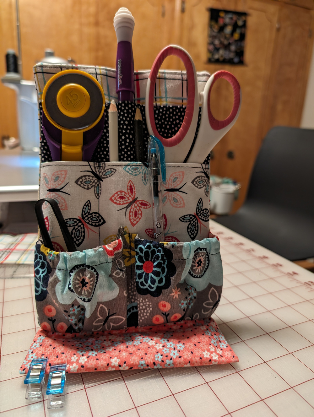 A fabric organizer holds scissors, rotary cutters, pens, and sewing clips, situated on a patterned surface with a sewing machine in the background.