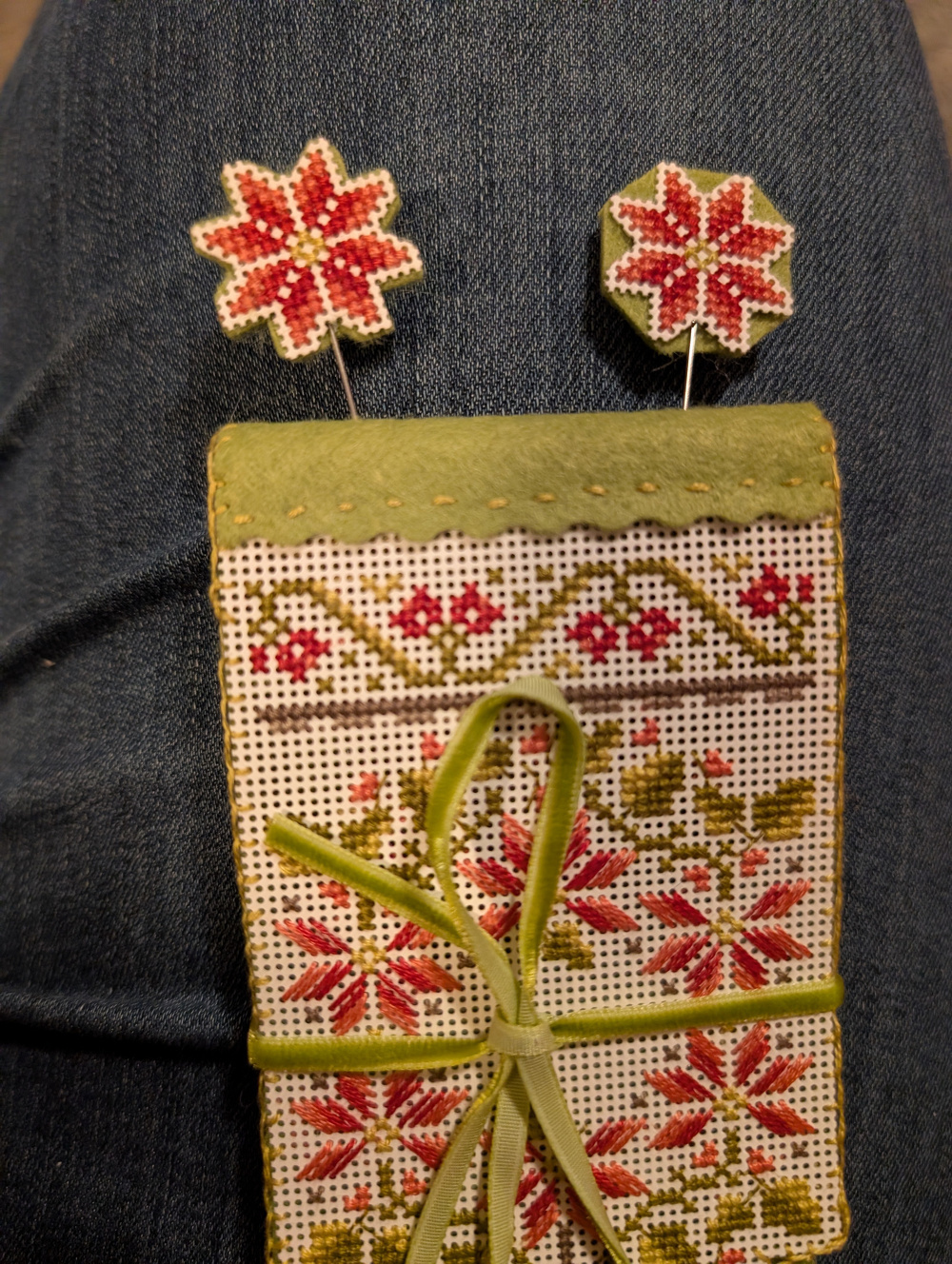 A handcrafted needle book with red and green floral embroidery is adorned with two matching decorative pins.