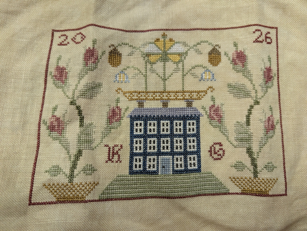 A cross-stitched design features a blue house surrounded by floral patterns, initials K G, and the numbers 20 and 26.