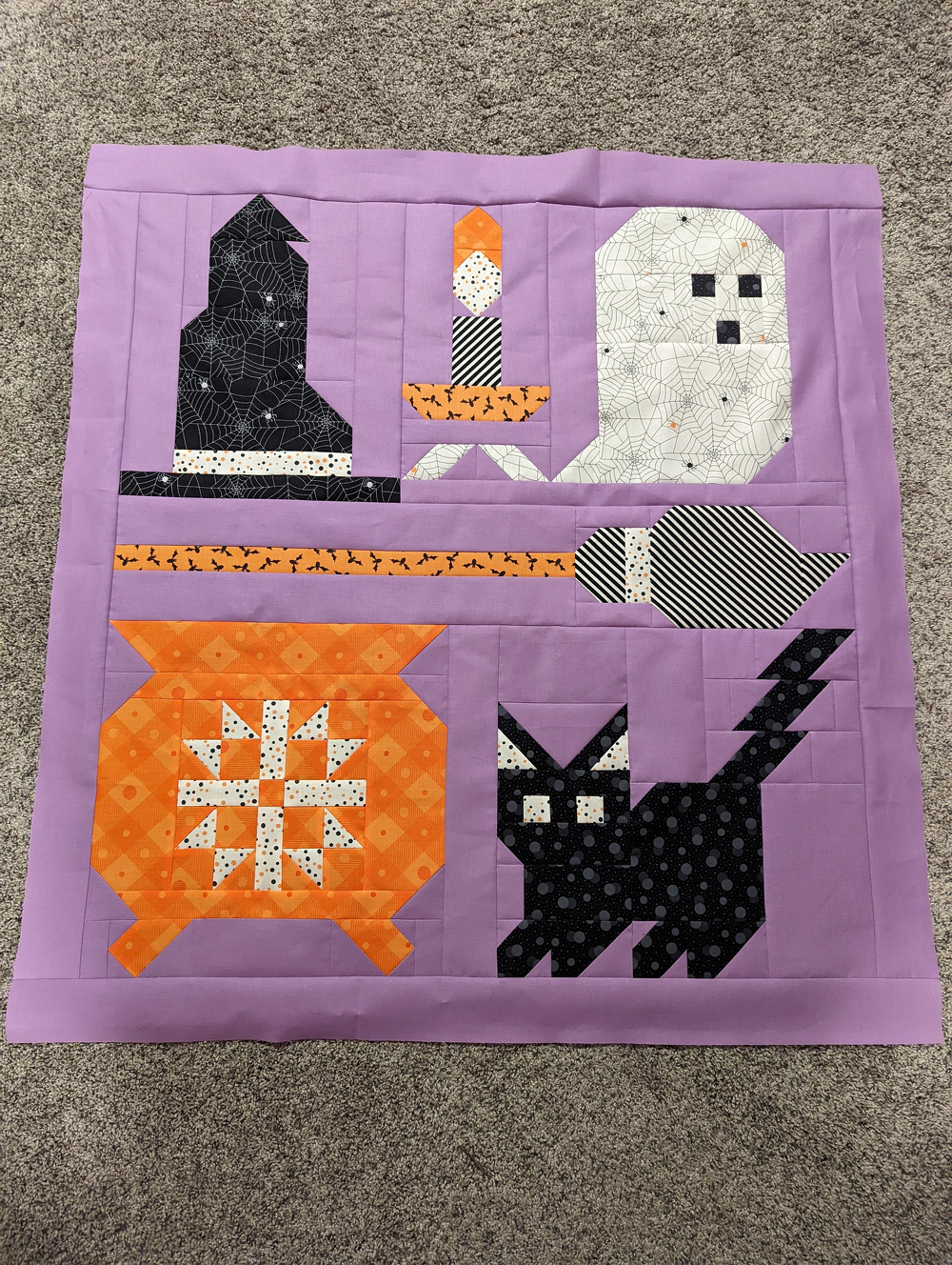 A quilt features Halloween-themed patchwork designs, including a witch's hat, candle, ghost, broom, cauldron, and a black cat, set against a purple background.
