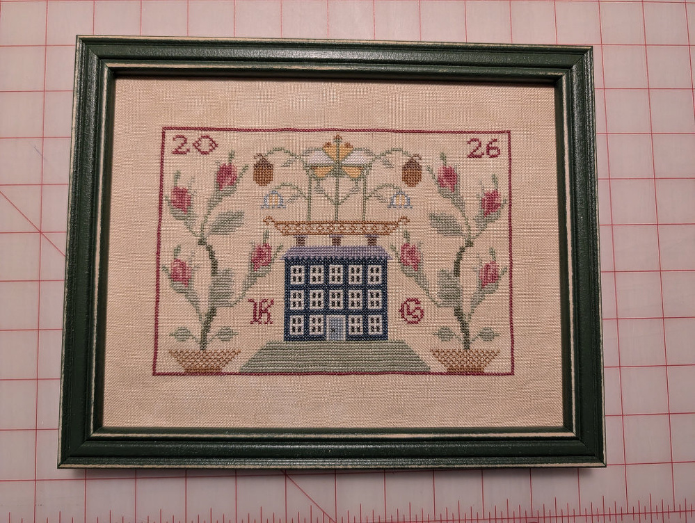 A framed cross-stitch artwork features a blue house centered between floral designs, with the year 2026 and the initials K and G stitched on either side.