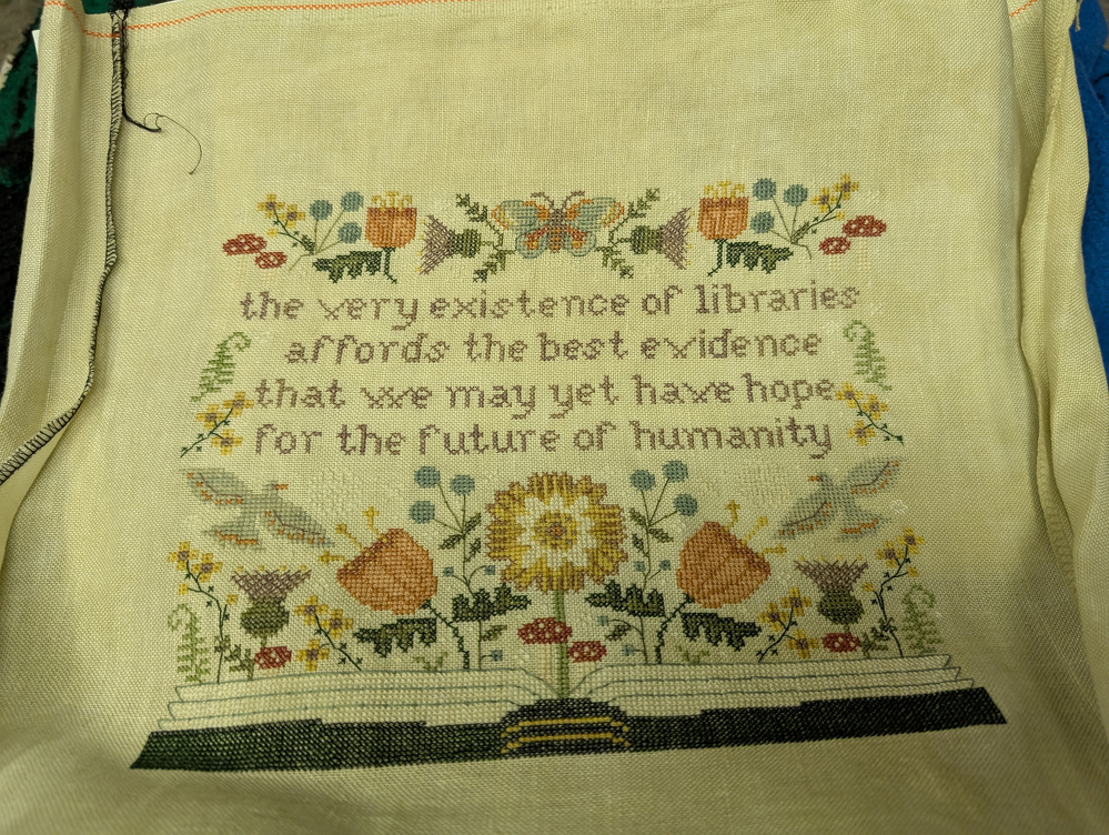 A cross-stitched piece features a quote about libraries surrounded by flowers and an open book.