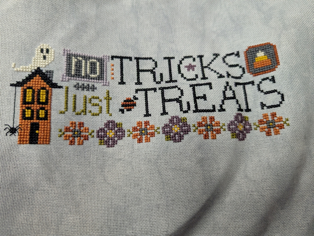 A cross-stitch design features a Halloween theme with a haunted house, ghost, and the text No Tricks, Just Treats adorned with flowers and festive icons.