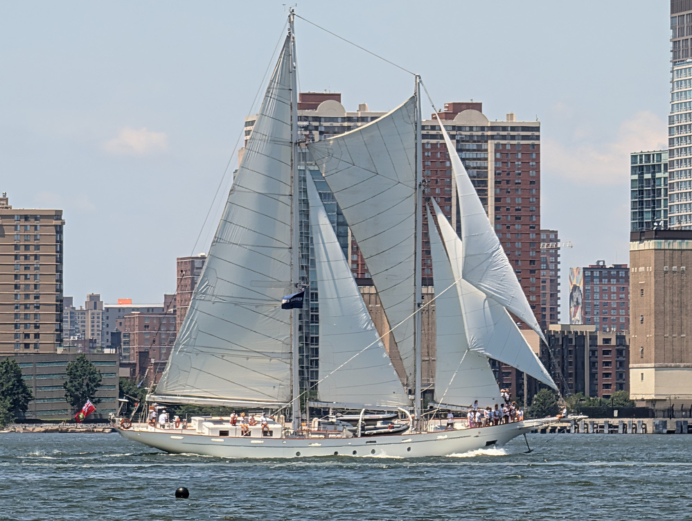 A sailing yacht with white sails is cruising on a body of water near a city with tall buildings in the background.