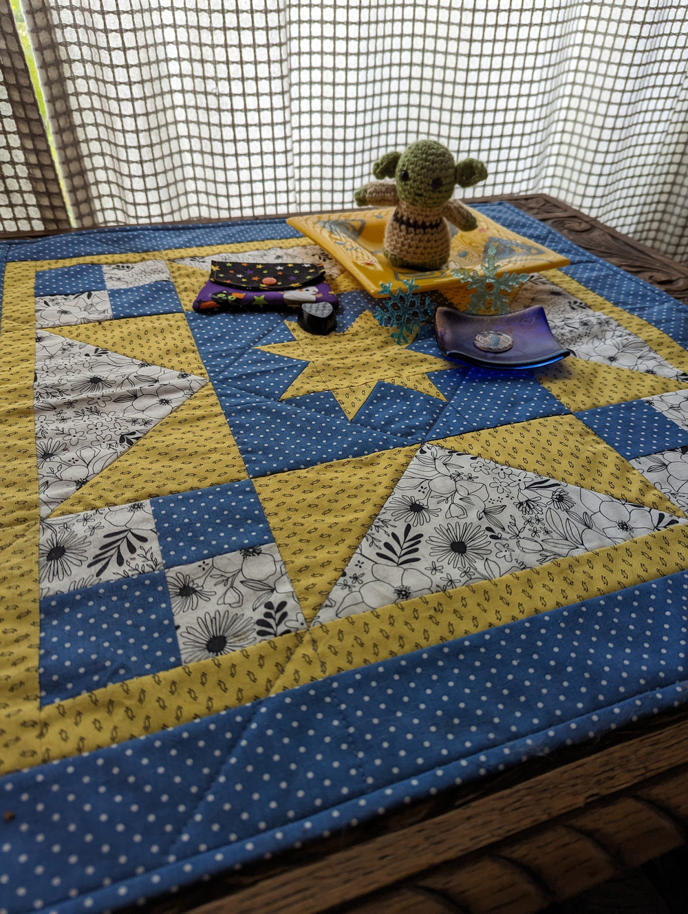 A quilted table mat with blue, yellow, and floral patterns is adorned with crochet items, including a small green figure, and various sewing accessories.