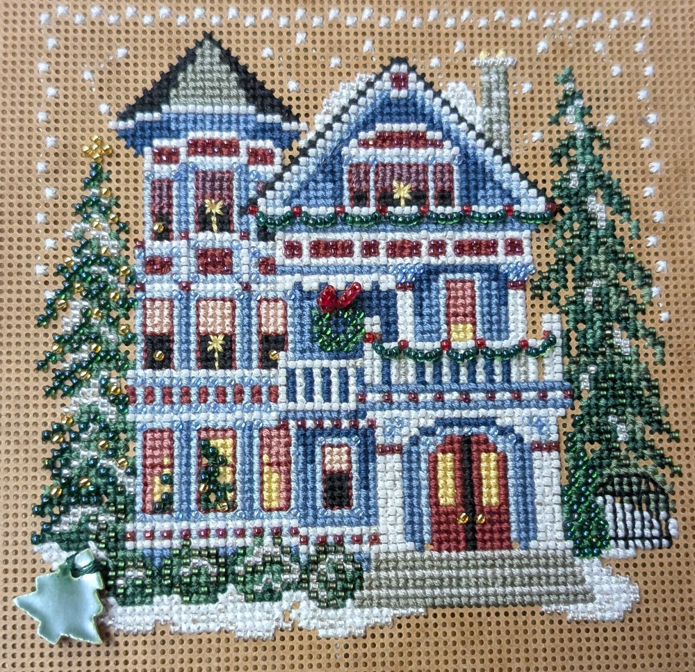 A cross-stitched picture depicts a festive, snow-covered house adorned with Christmas decorations and surrounded by trees.