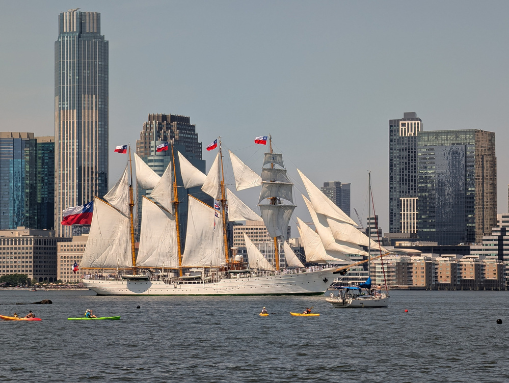 A large white sailing ship with multiple sails and flags is sailing on the water near a city skyline with kayakers nearby.