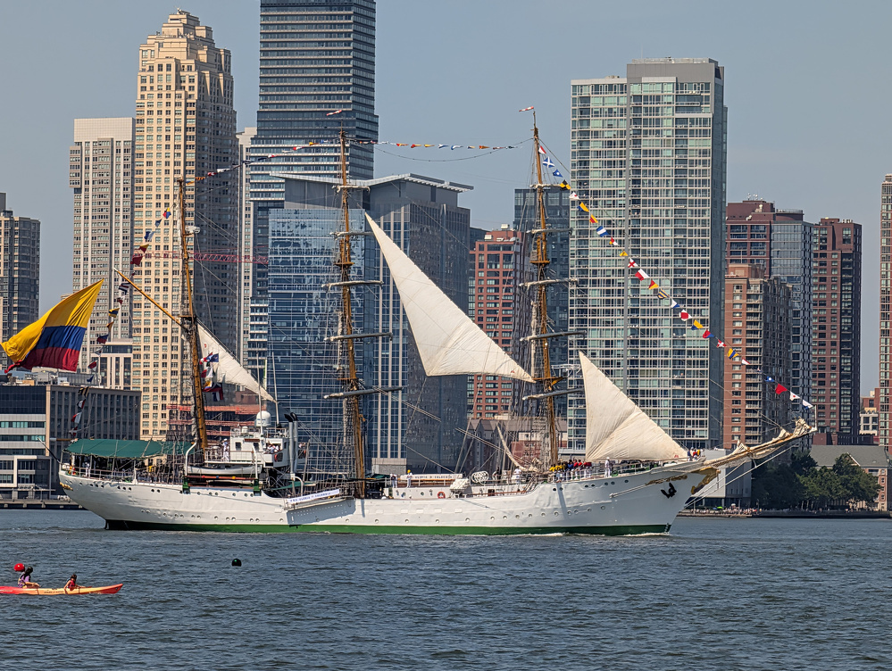 A large white sailing ship with multiple masts and colorful flags is floating on the water in front of a city skyline with tall buildings.