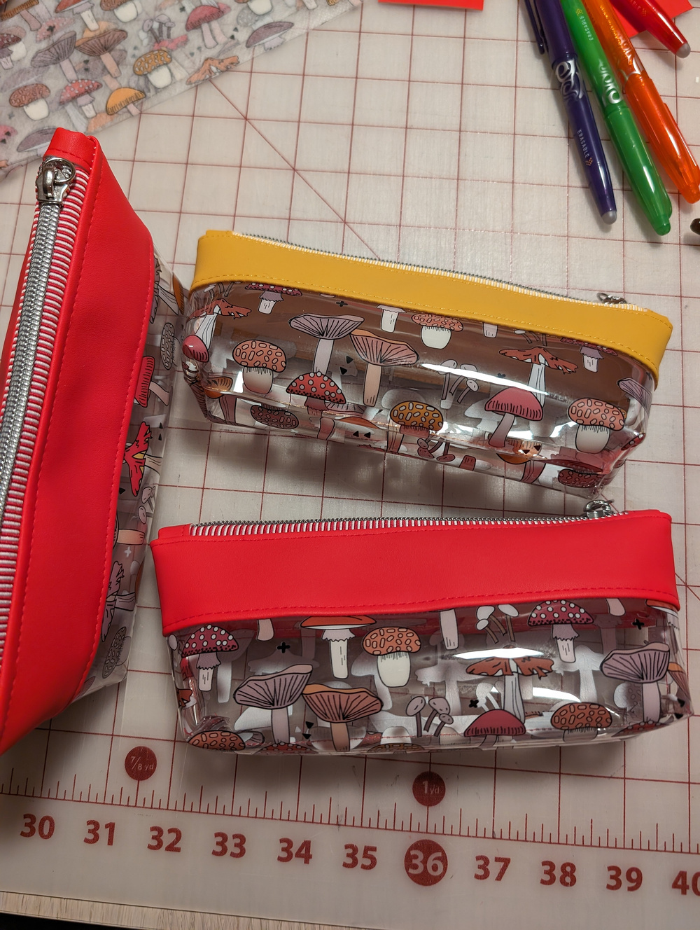 Three colorful pencil cases featuring mushroom patterns are arranged on a cutting mat next to colorful pens.