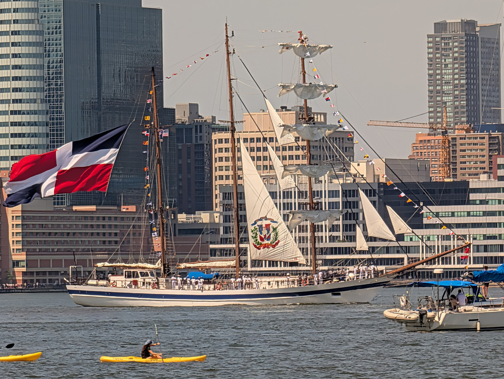 A large sailing ship with a Dominican Republic flag and emblem on its sails is docked near a city waterfront, while people paddle kayaks in the water nearby.
